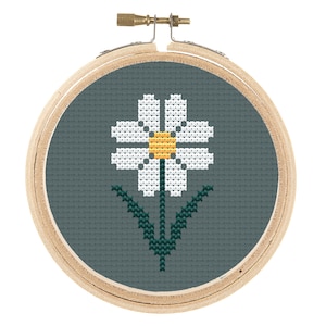 May include: A white daisy with a yellow center is embroidered on a teal fabric in a wooden embroidery hoop.