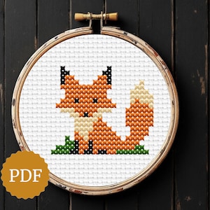 May include: A cross-stitch pattern of a fox in an embroidery hoop. The fox is orange, cream, and black, sitting on green grass. The hoop is made of wood. The PDF logo is in the bottom left corner.