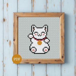 Peut inclure: Un motif de point de croix représentant un chat porte-bonheur blanc avec des contours noirs, des oreilles roses et un collier rouge avec une clochette dorée. Le motif est centré sur une toile à grille blanche dans un cadre en bois clair.