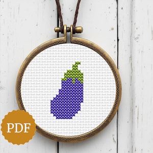 May include: A cross-stitch pendant featuring a purple eggplant with a green stem, set in a round, bronze-colored frame. The pendant hangs from a brown cord. The image includes a PDF icon.
