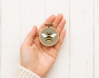 Può includere: Un piccolo telaio da ricamo circolare in legno contenente un disegno a punto croce di un'ape. L'ape presenta strisce orizzontali nere e gialle sul corpo e ali bianche, ricamate su uno sfondo di tessuto beige chiaro.