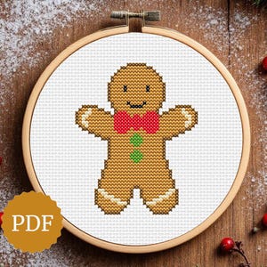 May include: Cross-stitch pattern of a gingerbread man with a red bow tie and green buttons, displayed in a wooden embroidery hoop. The background is a wooden surface with decorative elements, including red berries and greenery. The image includes a "PDF" icon.