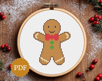 May include: Cross-stitch pattern of a gingerbread man with a red bow tie and green buttons, displayed in a wooden embroidery hoop. The background is a wooden surface with decorative elements, including red berries and greenery. The image includes a "PDF" icon.