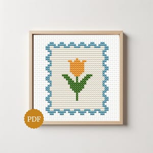 May include: A cross-stitch pattern featuring a central orange tulip with green leaves, enclosed within a scalloped blue border on a white grid background. The design is framed in a light wood square frame, with a gold circular PDF icon in the corner.