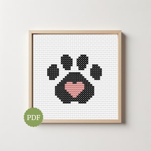 May include: A cross-stitch pattern featuring a black paw print design on a white grid background. The center of the paw pad contains a small, light pink heart shape. The entire design is framed in a light wood square frame with a green PDF icon.