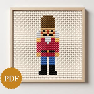 May include: A framed cross-stitch of a nutcracker soldier. The artwork features a brown-haired nutcracker with a red jacket, blue pants, and black boots. The background is a beige fabric-like texture. A PDF logo is in the lower left corner.