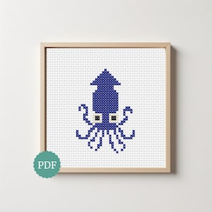 May include: A cross-stitch pattern featuring a dark blue squid centered on a white grid background, framed in a light wood square frame. A teal circular badge with the white text PDF is positioned in the bottom left corner of the image.