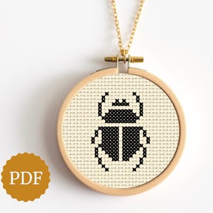 May include: A gold-colored necklace featuring a round pendant with a cross-stitch design of a black beetle on a cream-colored background. The pendant is set in a wooden embroidery hoop. The image also includes a PDF icon.