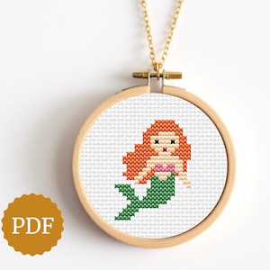 Può includere: Una collana con pendente a punto croce con un disegno di sirena. La sirena ha i capelli rossi, un top rosa e una coda verde, su sfondo bianco all'interno di un cerchio di legno. La collana ha una catena d'oro.