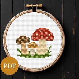 May include: Cross-stitch embroidery of three mushrooms in a wooden hoop. The mushrooms have red, brown, and tan caps with white spots, set against a white background. Green grass is at the base. The PDF logo is in the lower left.