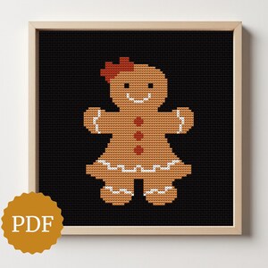 May include: A framed cross-stitch pattern of a gingerbread person on a black background. The gingerbread person is brown with white icing details, red bow, and red buttons. The frame is light brown, and a PDF logo is in the lower left corner.
