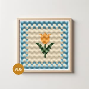 Può includere: Un motivo a punto croce quadrato in una cornice di legno chiaro con un tulipano arancione centrale con foglie verdi su sfondo crema, circondato da un bordo a scacchi blu e crema. È presente un badge circolare dorato con il testo PDF.