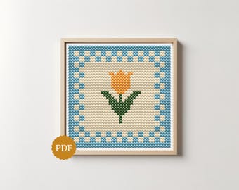 May include: A square cross-stitch pattern in a light wood frame featuring a central orange tulip with green leaves on a cream background, surrounded by a blue and cream checkered border. A gold circular badge with the text PDF is in the corner.