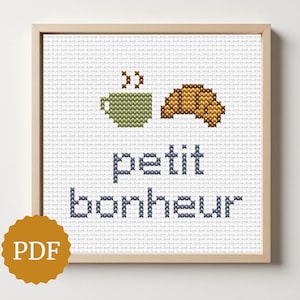 May include: Cross-stitch art in a wooden frame. The design features a green cup, a brown croissant, and the words "petit bonheur" in blue. A PDF logo is in the lower left corner.