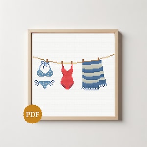 Può includere: Un motivo a punto croce incorniciato in legno chiaro raffigurante un bikini blu a pois, un costume intero rosso tinta unita e un telo mare a righe blu e bianche appesi a un filo da bucato con mollette di legno su sfondo a griglia bianca.