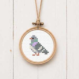 May include: A circular wooden embroidery hoop containing a cross-stitch pattern of a pigeon. The bird is depicted in shades of gray, white, purple, and green, with an orange beak and feet. The hoop is suspended by a thin, light brown twine loop.
