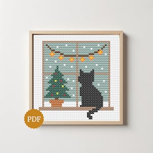 May include: A cross-stitch pattern in a light wood frame depicting a black cat sitting on a windowsill. Outside the window, a green Christmas tree with colourful ornaments stands in the snow, and a string of orange lights hangs above.