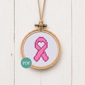 Puede incluir: Un bastidor de bordado circular de madera que contiene un patrón de punto de cruz de una cinta de concienciación rosa con un recorte en forma de corazón en el centro. La tela es blanca con un patrón de cuadrícula y un lazo de cuerda beige.