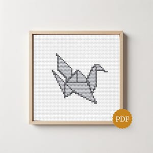 Op de afbeelding: Een kruissteekpatroon met een grijze origami kraanvogel gecentreerd op een witte rasterachtergrond, ingelijst in een vierkante lichthouten lijst. Een kleine, ronde goudkleurige badge met de letters PDF bevindt zich in de rechterbenedenhoek.