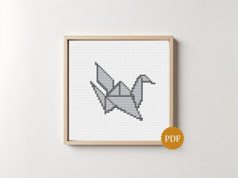Op de afbeelding: Een kruissteekpatroon met een grijze origami kraanvogel gecentreerd op een witte rasterachtergrond, ingelijst in een vierkante lichthouten lijst. Een kleine, ronde goudkleurige badge met de letters PDF bevindt zich in de rechterbenedenhoek.