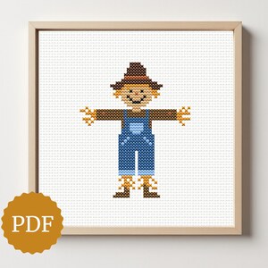 May include: Cross-stitch art of a smiling scarecrow in a wooden frame. The scarecrow wears a brown hat, brown shirt, blue overalls, and straw-like arms and legs. The image includes a PDF logo in a gold circle.