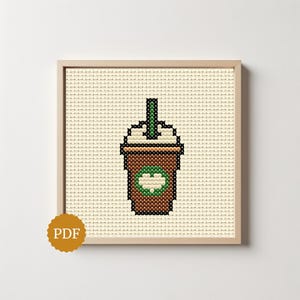 Op de afbeelding: Een kruissteekpatroon met een bruine koffiebeker met witte slagroom en een groen rietje, gecentreerd op een lichtbeige stoffen raster. De beker toont een groen cirkelvormig embleem met een wit hart in het midden, ingelijst in licht hout.