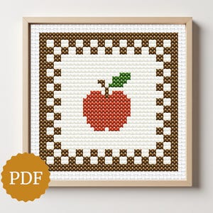 May include: Cross-stitch artwork of a red apple with a green leaf, framed in a wooden frame. The apple is centered on a white background, surrounded by a brown and white checkered border. The image includes a PDF icon.