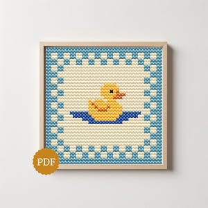 Peut inclure: Un motif de point de croix représentant un canard en caoutchouc jaune avec un bec orange flottant sur une petite mare bleue. Le motif est encadré par une bordure à carreaux alternant des carrés bleus et crème, le tout dans un cadre en bois clair.