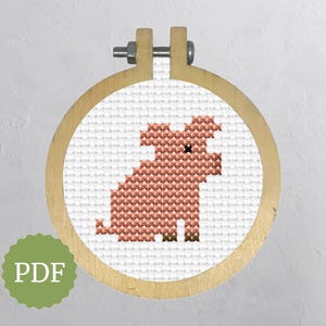 May include: Cross-stitch piglet design in a wooden embroidery hoop. The pig is rendered in shades of pink and brown against a white background. The hoop is secured with a metal screw and wooden frame. A green PDF logo is in the lower left.