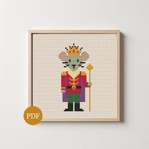 May include: A cross-stitch pattern featuring a gray mouse dressed as a king, wearing a yellow crown, a red jacket with gold details, a purple cape, and green trousers, holding a gold scepter, framed in a light wood square frame.