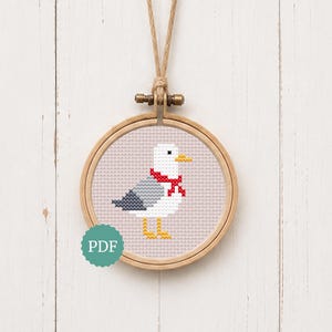 May include: A cross-stitch pattern of a seagull wearing a red scarf, displayed in a round wooden embroidery hoop. The seagull has a white head and body, grey wings, a yellow beak, and yellow feet. A teal circular icon with the text PDF is visible.