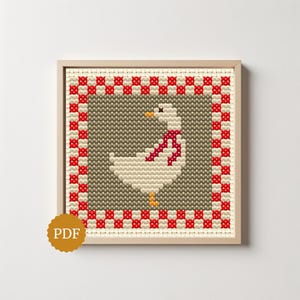 Peut inclure: Un motif de point de croix carré représentant une oie blanche avec un bec et des pattes orange, portant une écharpe rouge. L'oie est centrée sur un fond gris, encadrée par une bordure à carreaux rouges et blancs, dans un cadre en bois clair.