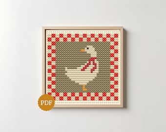 May include: A square cross-stitch pattern featuring a white goose with an orange beak and feet, wearing a red scarf. The goose is centered on a gray background, framed by a red and white checkered border, all enclosed in a light wood frame.