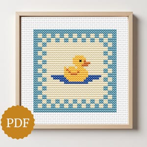 May include: A framed cross-stitch pattern featuring a yellow rubber duck floating on blue water. The duck is centered within a square design with a blue border. The frame is light brown, and a PDF icon is in the lower left corner.