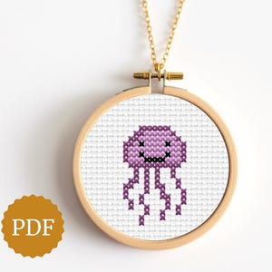 Puede incluir: Un collar con colgante de punto de cruz con un diseño de medusa morada sonriente. El diseño está enmarcado en un aro de bordado de madera y cuelga de una cadena dorada. Un accesorio único y hecho a mano.