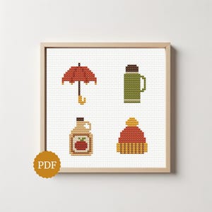 May include: A cross-stitch pattern featuring four icons: a red umbrella, a green thermos, a jug of maple syrup with a red apple label, and a red and yellow knit beanie. The design is displayed in a light wood frame with a PDF icon in the corner.