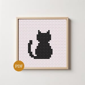 Peut inclure: Un modèle de point de croix représentant la silhouette d'un chat noir remplie de pois blancs sur un fond à grille rose pâle, encadré dans un cadre carré en bois clair, avec un badge orange circulaire marqué PDF dans le coin inférieur.