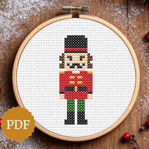 Puede incluir: Patrón de punto de cruz de un soldado de juguete en un aro de bordado de madera. El soldado viste un abrigo rojo con botones dorados, botas negras y un sombrero negro. El fondo es una superficie de madera con decoraciones y el texto "PDF".