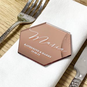 May include: A rose gold hexagonal acrylic place card with the name "Maria" written in white cursive script. The text "Katherine & Harry 19.08.23" is also written in white below the name.