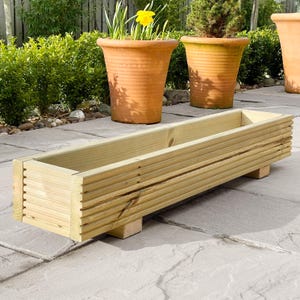 May include: A long, rectangular wooden planter box with a slatted design. The planter is sitting on a patio with stone paving slabs. The planter is empty.