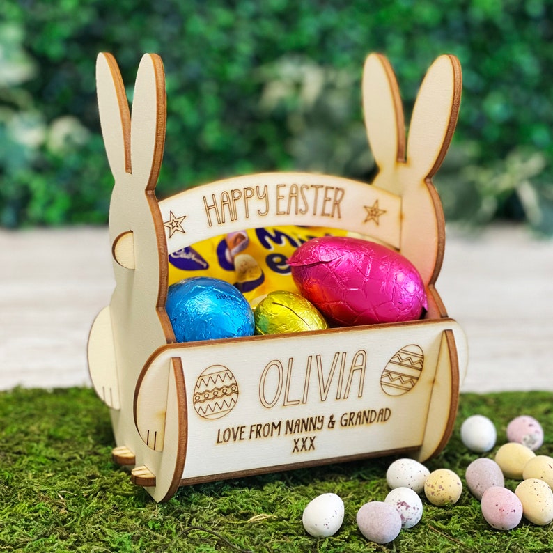 May include: A wooden Easter basket shaped like a bunny, filled with foil-wrapped chocolate eggs. The basket has the words "Happy Easter" and a name engraved on it. Additional decorative eggs are scattered around the basket.