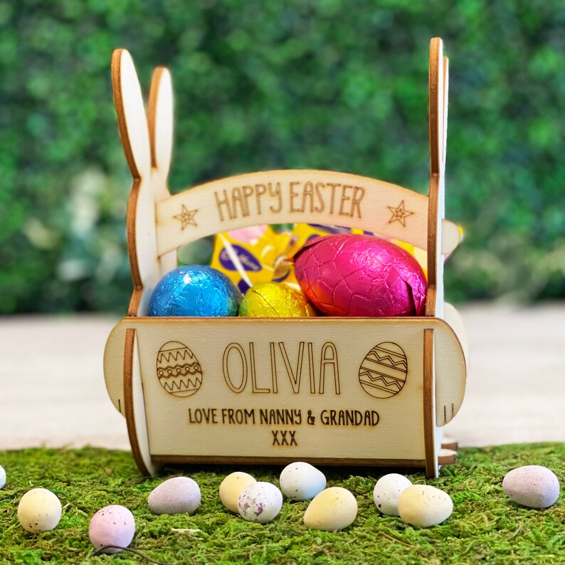 May include: A wooden Easter basket shaped like a bunny, filled with foil-wrapped chocolate eggs. The basket has the words "HAPPY EASTER" and "OLIVIA" engraved on it, along with a message from family. The basket sits on a bed of green moss, surrounded by Easter eggs.
