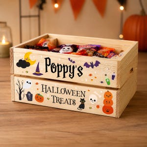 May include: A light-coloured wooden crate filled with assorted sweets, featuring the text Poppy's HALLOWEEN TREATS. The crate is decorated with colourful illustrations of a yellow moon, purple witch hat, bats, spider web, ghost, tombstone, pumpkins, and a black cat.