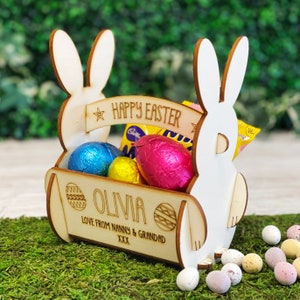May include: A wooden Easter basket shaped like a bunny, filled with foil-wrapped chocolate eggs. The basket has the text "HAPPY EASTER" and the name "OLIVIA". The basket is sitting on a bed of green moss, with scattered Easter eggs.