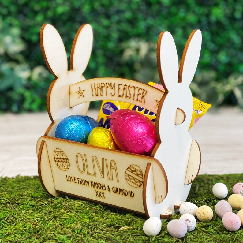 May include: A wooden Easter basket shaped like a bunny, filled with foil-wrapped chocolate eggs. The basket has the text "HAPPY EASTER" and the name "OLIVIA". The basket is sitting on a bed of green moss, with scattered Easter eggs.