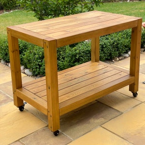 May include: A wooden table with two shelves. The table is made of natural wood and has a rustic look. It has wheels on the bottom for easy mobility.