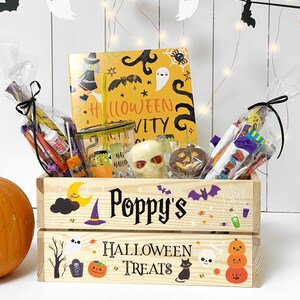 May include: A wooden crate labelled Poppy's Halloween Treats, decorated with pumpkins, ghosts, and bats. It contains a yellow activity book, a clear tumbler with a straw, a skull-shaped treat, a chocolate pumpkin, and assorted wrapped sweets.