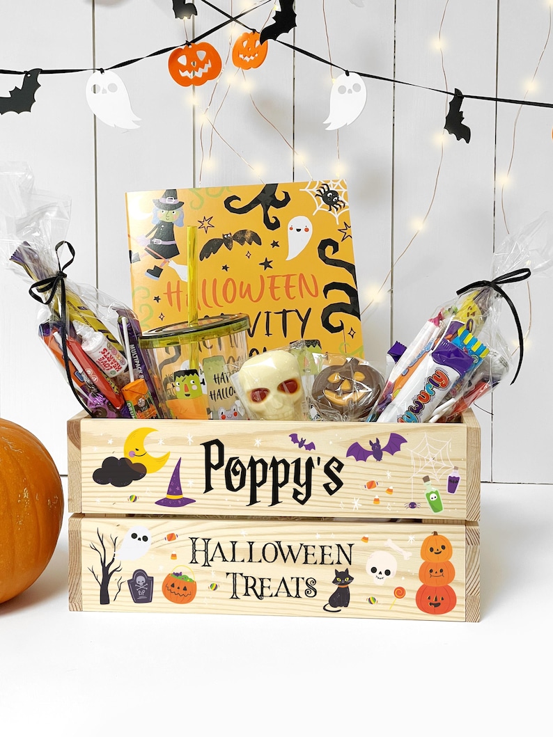 May include: A wooden crate labelled Poppy's Halloween Treats, decorated with pumpkins, ghosts, and bats. It contains a yellow activity book, a clear tumbler with a straw, a skull-shaped treat, a chocolate pumpkin, and assorted wrapped sweets.