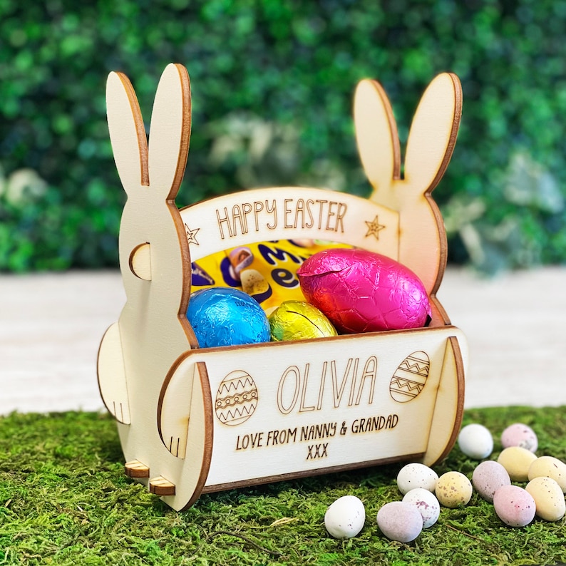 May include: A wooden Easter basket shaped like a bunny, filled with foil-wrapped chocolate eggs. The basket has the words "HAPPY EASTER" and "OLIVIA" engraved on it, along with the message "LOVE FROM NANNY & GRANDAD XXX". The basket is on a bed of green moss.