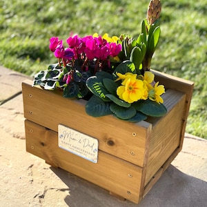 May include: A wooden planter box with a personalised sign that reads "Mum & Dad Happy Anniversary Lots of Love Happy 25th". The planter is filled with pink and yellow flowers and green foliage.